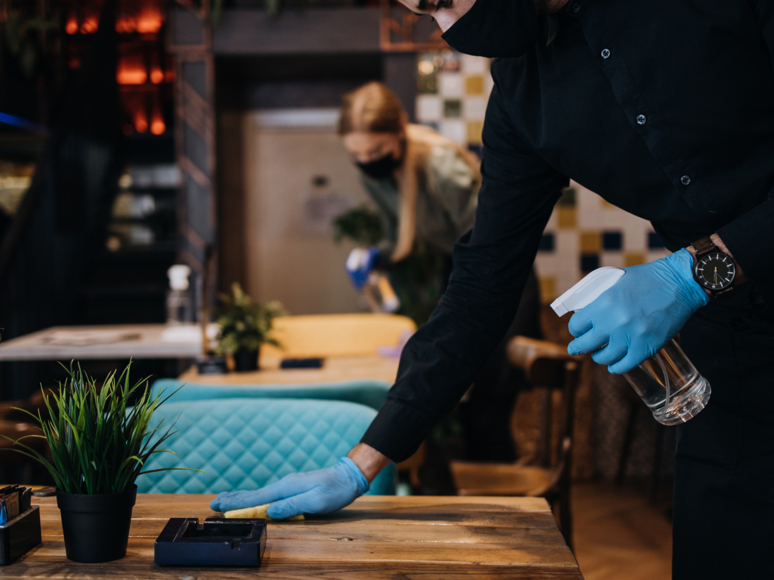 Medewerker met mondkapje en blauwe handschoenen spuit en poetst een houten restauranttafel.