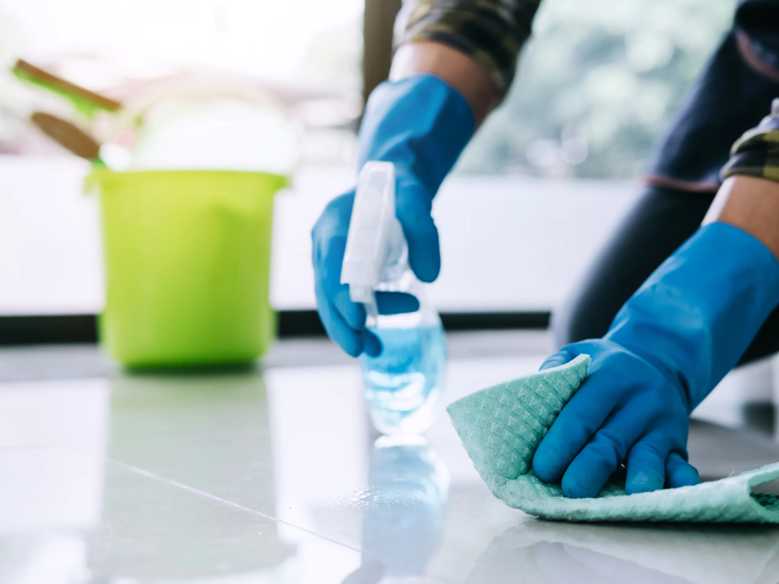 Persoon met werkhandschoenen reinigt een tegelvloer met een microvezeldoek en spray.